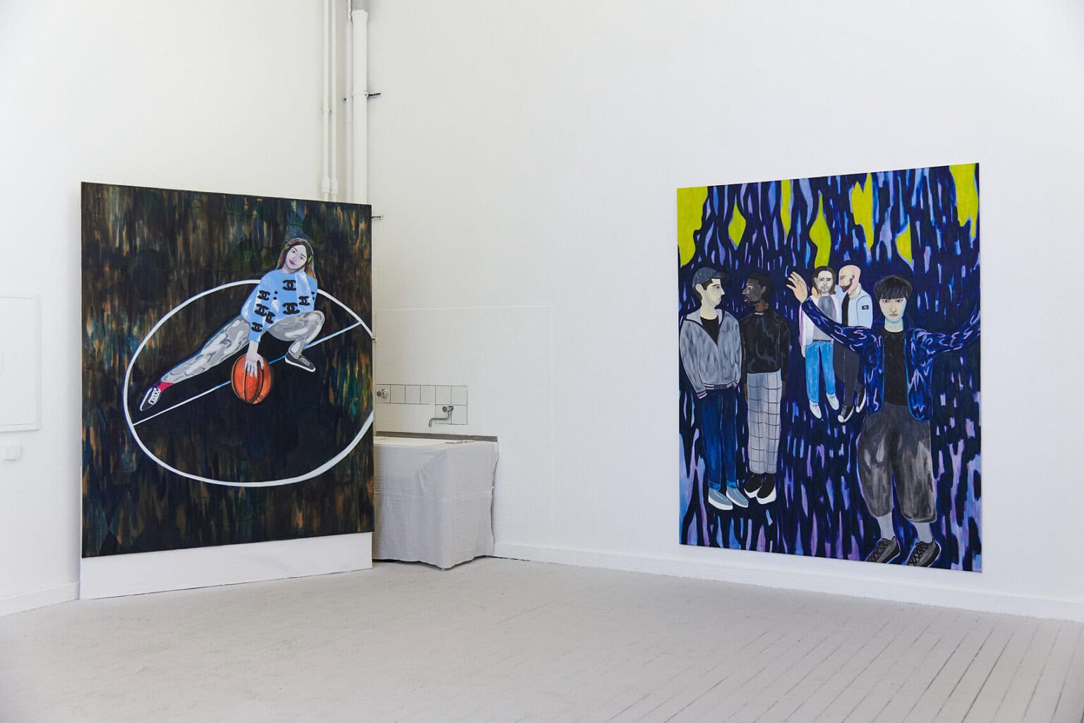 Foto: Ein Raum mit weißen Wänden und grauem Dielenboden. An einer der Wände hängt eine großformatige Leinwand im Hochformat, in der Ecke des Raumes auf dem Boden steht eine weitere.