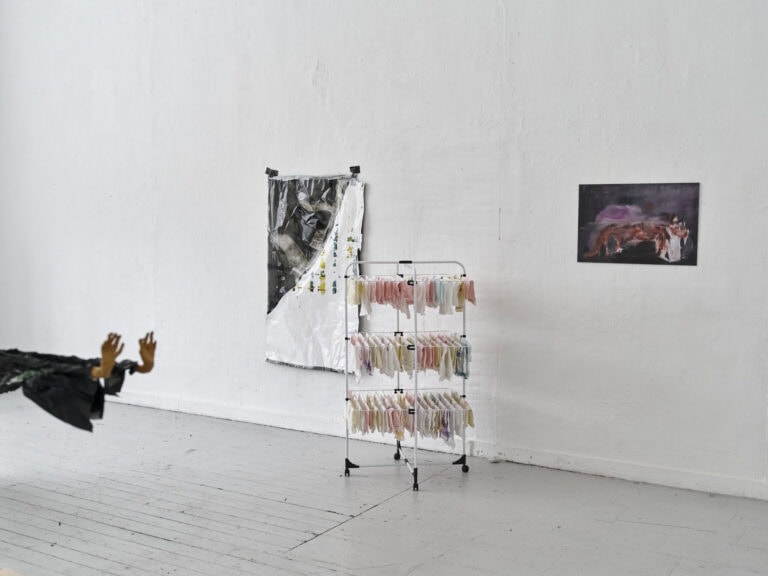 Foto: Aufnahme eines Raumes mit weißen Wänden und grauem Dielenboden. An den Wänden hängen flache Objekte. Im Raum stehen Objekte.