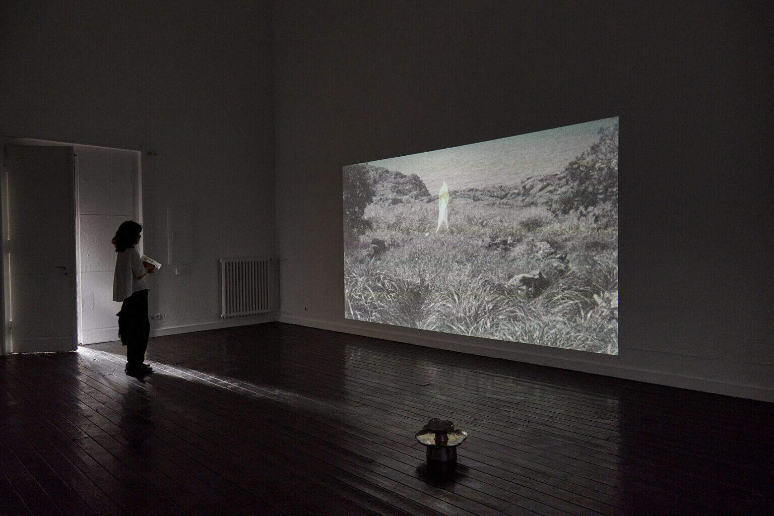 Foto: Ein abgedunkelter Raum mit Dielenboden. An die Wand wird eine großformatige Projektion geworfen.Auf dem Boden steht ein Objekt und im Raum steht ein Mensch. Die Tür des Raumes ist einen spalt weit geöffnet und Licht fällt hinein.