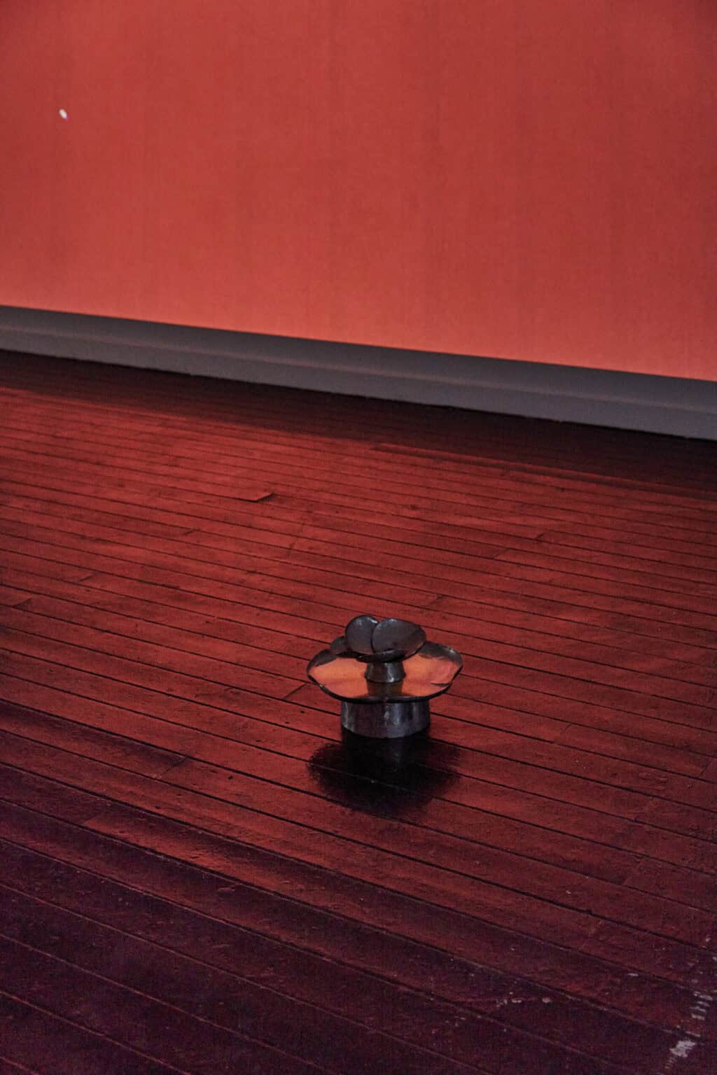 Foto: Ein abgedunkelter Raum mit Dielenboden. Auf dem Boden steht ein Objekt. Der Boden und das Objekt reflektiert rotes Licht, das an die Wand geworfen wird.