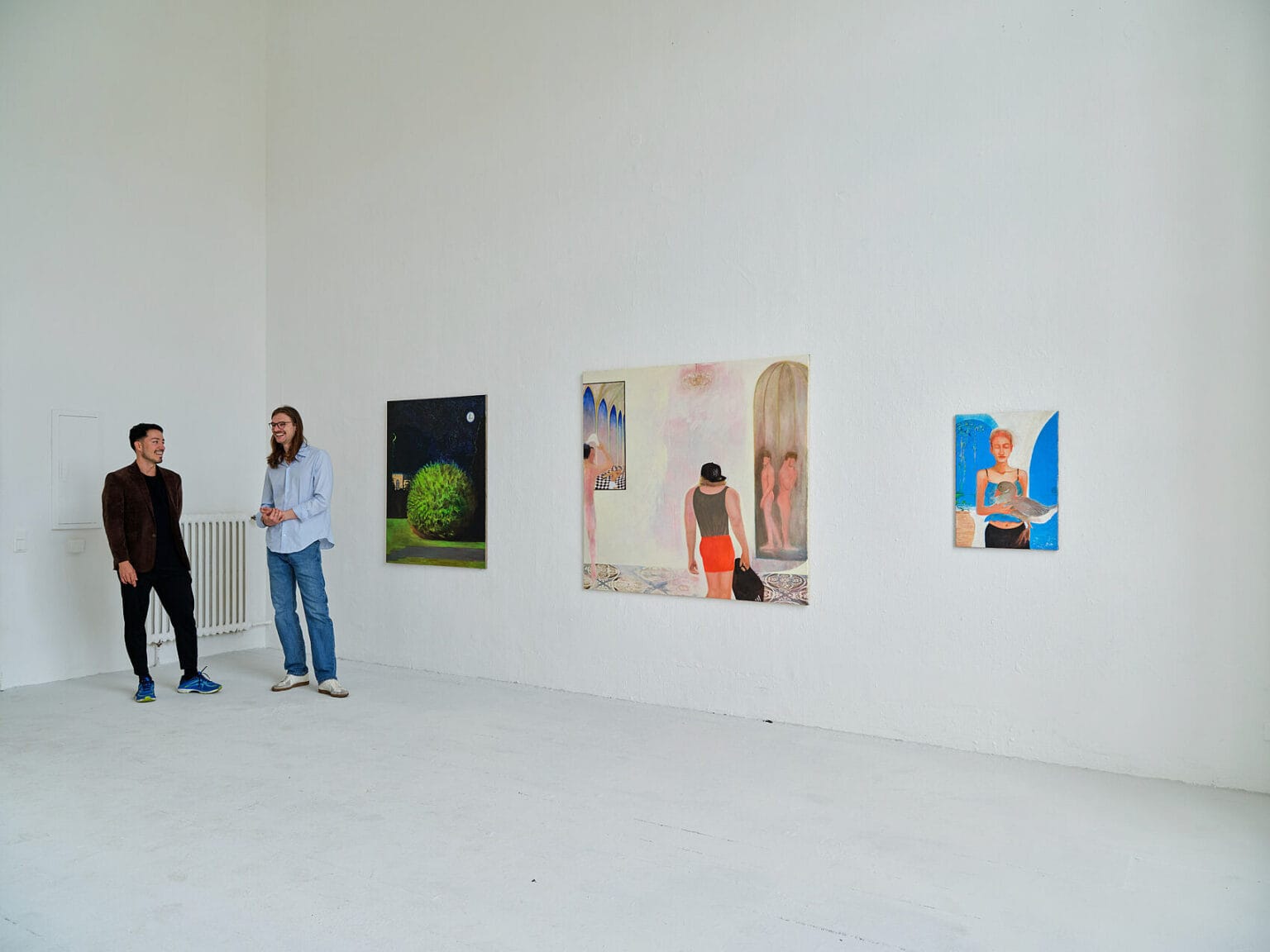 Foto: Raum mit weißen Wänden und hellgrauem Boden. An der Wand Leinwandarbeiten im Mittel- bis Großformat. Im Raum stehen zwei Menschen.