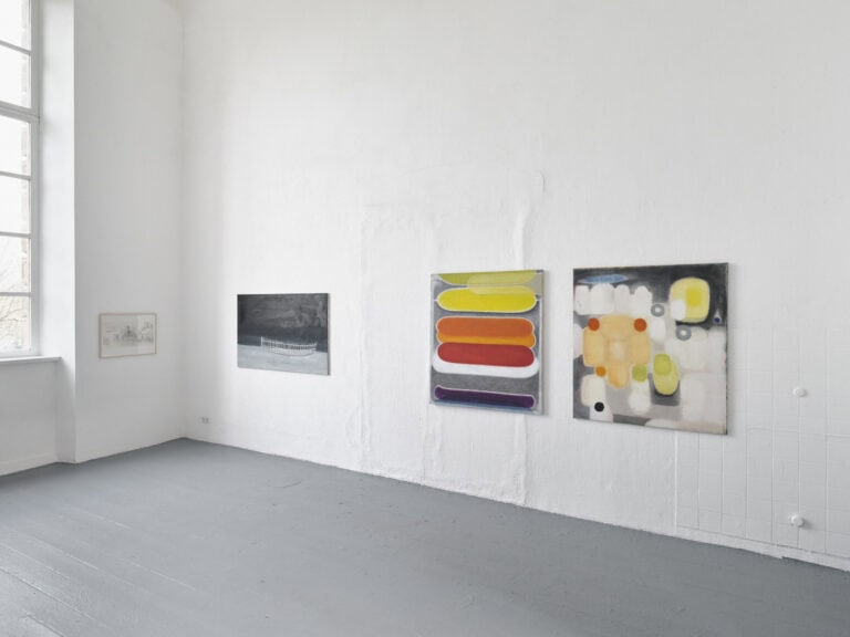 Foto: Aufnahme eines Raumes mit weißen Wänden, grauem Dielenboden und einem Fenster. An den Wänden hängen Malereien.