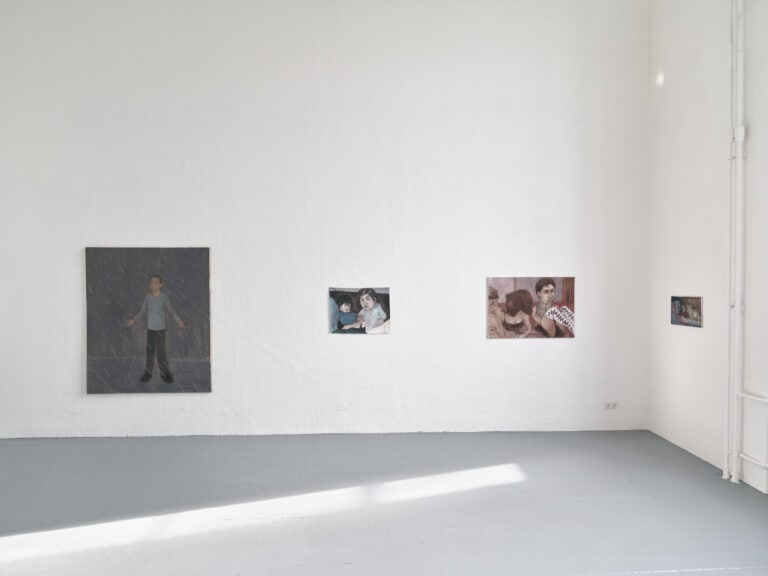 Foto: Aufnahme eines Raumes mit weißen Wänden und grauem Dielenboden. Ein Lichtreflex auf dem Boden. An der Wand hängen Bilder im Klein- und Mittelformat. zweiflüglige, geöffnete Tür. Hinter der Tür liegt ein großes Fenster, durch das Sonnenlicht fällt. Über der Tür hängt eine Malerei im Mittelformat.