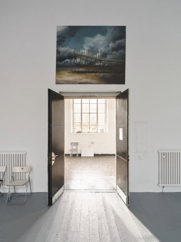 Foto: Aufnahme eines Raumes. Eine zweiflüglige, geöffnete Tür. Hinter der Tür liegt ein großes Fenster, durch das Sonnenlicht auf grauen Dielenboden fällt. Über der Tür hängt eine Malerei im Mittelformat.