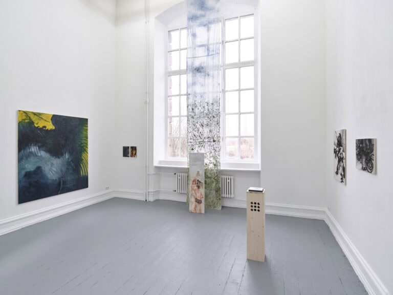 Foto: Aufnahme eines Raumes mit weißen Wänden, grauen Dielenboden und einem großen Fenster. Vor dem Fenster ist ein bemaltes, halbtransparentes Textil installiert. An der Wand befinden sich Malereien in Klein-, Mittel- und Großformaten. Im Raum befinden sich Objekte, zum Teil auf dem Boden, zum Teil auf Sockeln.