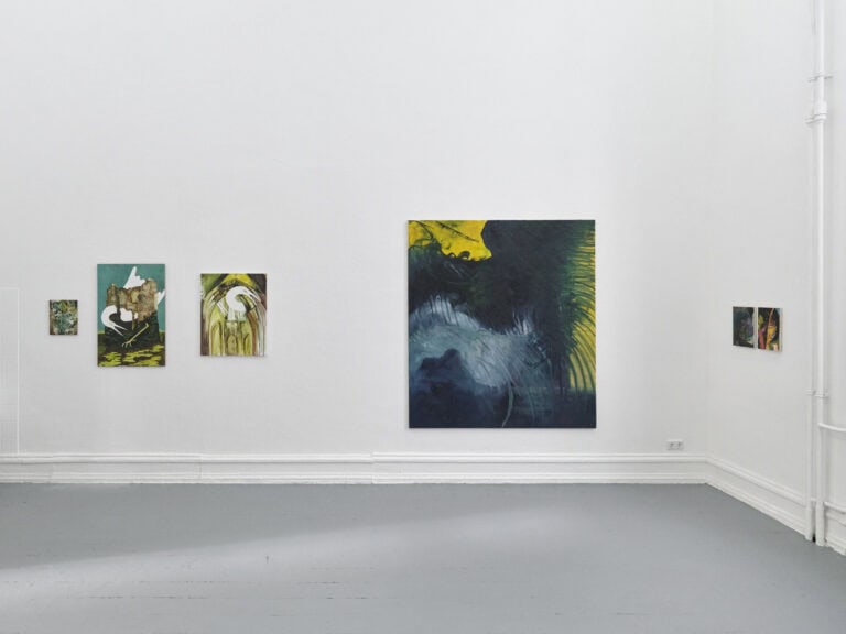 Foto: Aufnahme eines Raumes mit weißen Wänden und einem grauen Dielenboden. An der Wand befinden sich Malereien in Klein-, Mittel- und Großformaten.