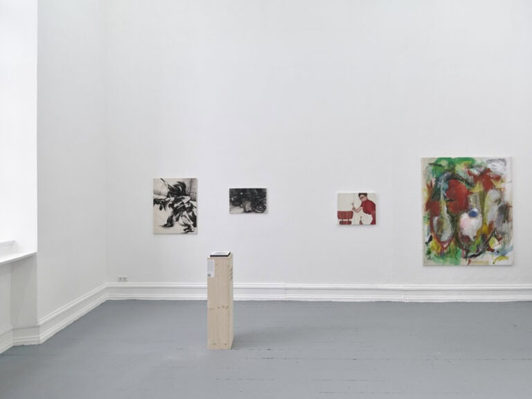 Foto: Aufnahme eines Raumes mit weißen Wänden und einem grauen Dielenboden. An den Wänden hängen Malereien in Klein- und Mittelformaten. Im Raum befindet sich ein Objekt auf einem Sockel.