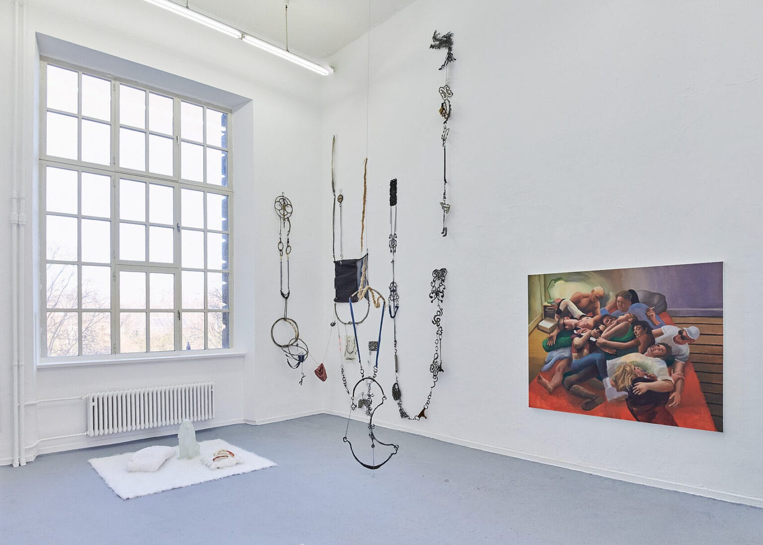 Foto: Ansicht eines Raumes mit weißen Wänden, grauem Boden und einem großen Fenster. An der Wand befindet sich eine großformatige Malerei und eine Konstruktion aus unterschiedlichen Materialien, die auf unterschiedlichen Höhen an der Wand befestigt sind und in Ketten herunterhängen. Auf dem Boden liegt eine Gruppe aus Objekten.