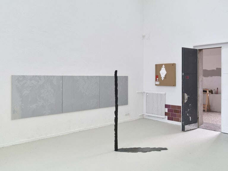 Foto: Aufnahme eines Raumes mit weißen Wänden, grau-grünem Boden und einer Tür. An den Wänden hängen Objekte und Malereien, auf dem Boden stehen und liegen Objekte.