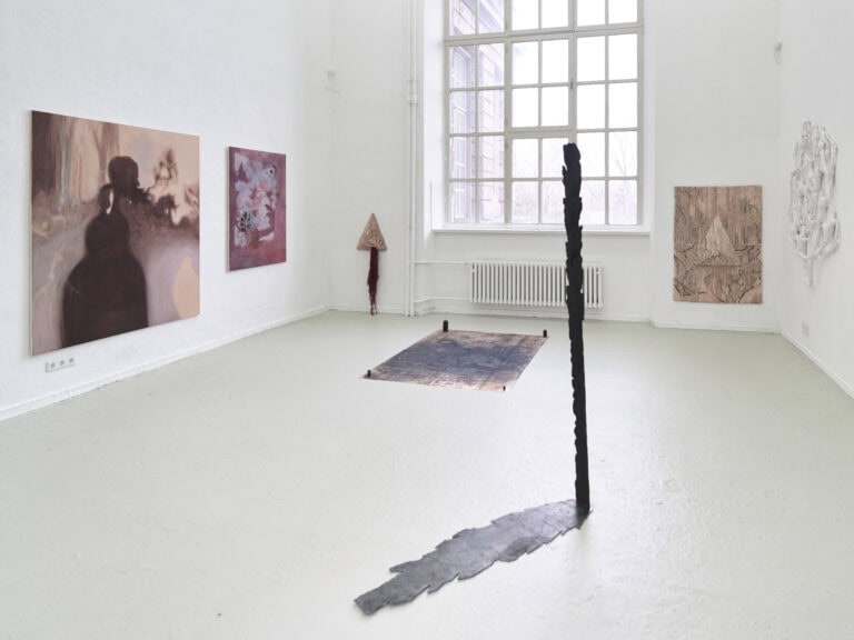 Foto: Aufnahme eines Raumes mit weißen Wänden, grau-grünem Boden und einem großen Fenster. An den Wänden hängen Objekte und Malereien, auf dem Boden stehen und liegen Objekte.