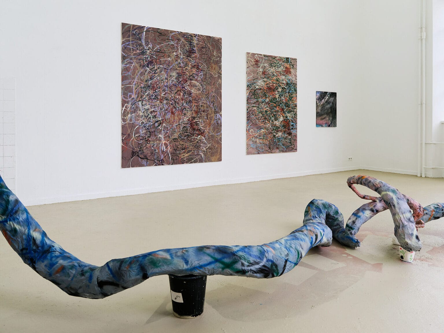 Foto: Ansicht eines Raumes mit weißen Wänden und beigem Boden. An der Wand des Raumes befinden sich drei groß- bis mittelformatige Malereien. Über den Boden des Raumes erstreckt sich ein längliches, buntes Objekt.