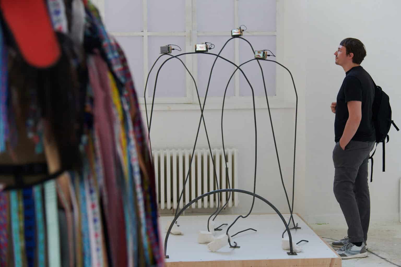 Foto: Ansicht eines Raumes mit weißen Wänden und einem Fenster. Im Raum befindet sich ein Post, auf dem sich Objekte aus unterschiedlichen Materialien befinden. Davor steht eine Person.