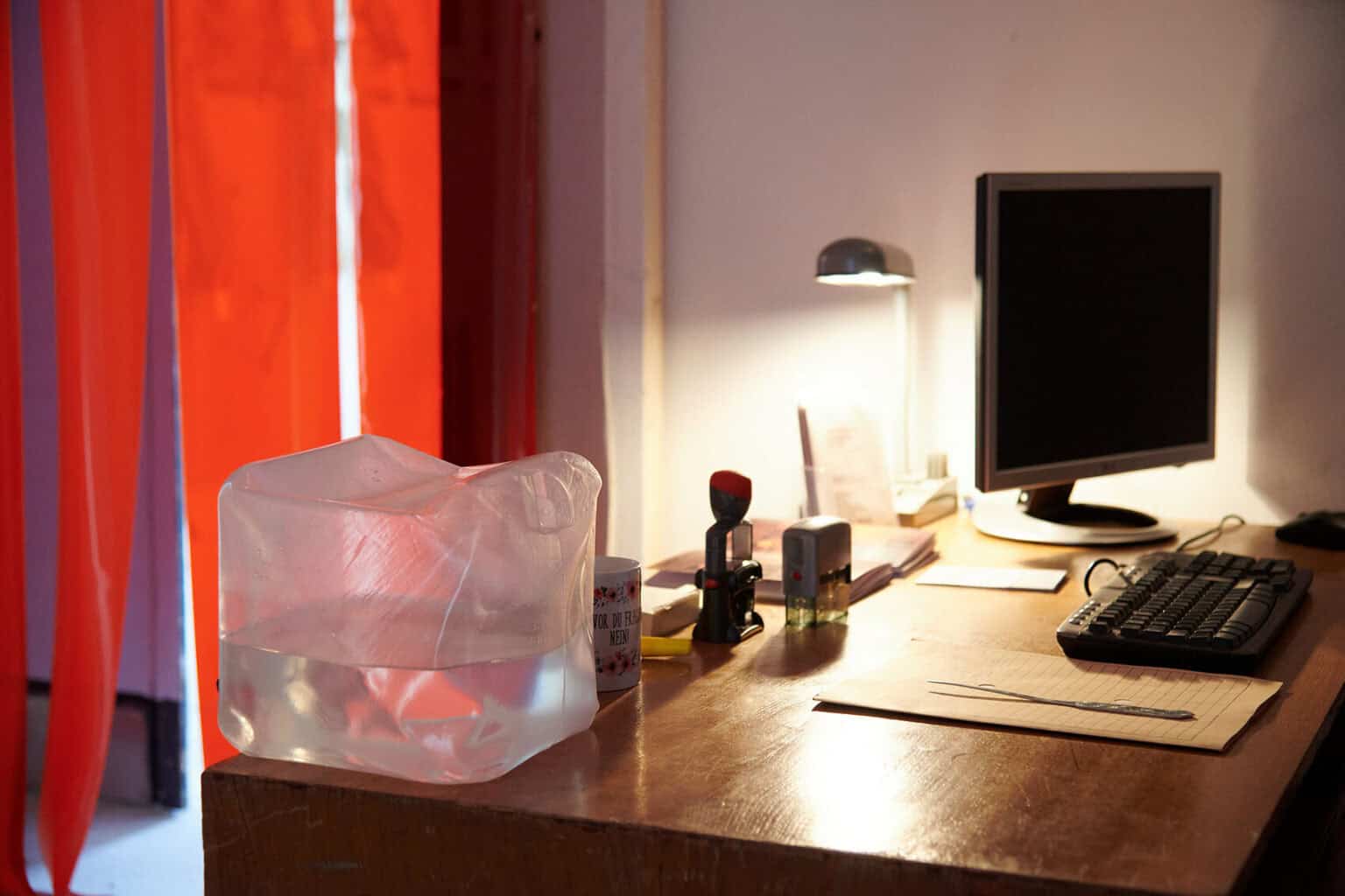 Foto: Ansicht eines Schreibtischs mit Büroeinrichtung, darunter eine Tischlampe, eine Tasse, ein Bildschirm, Stempel usw. Im Hintergrund ein Türrahmen, in dem ein roter Vorhang hängt.