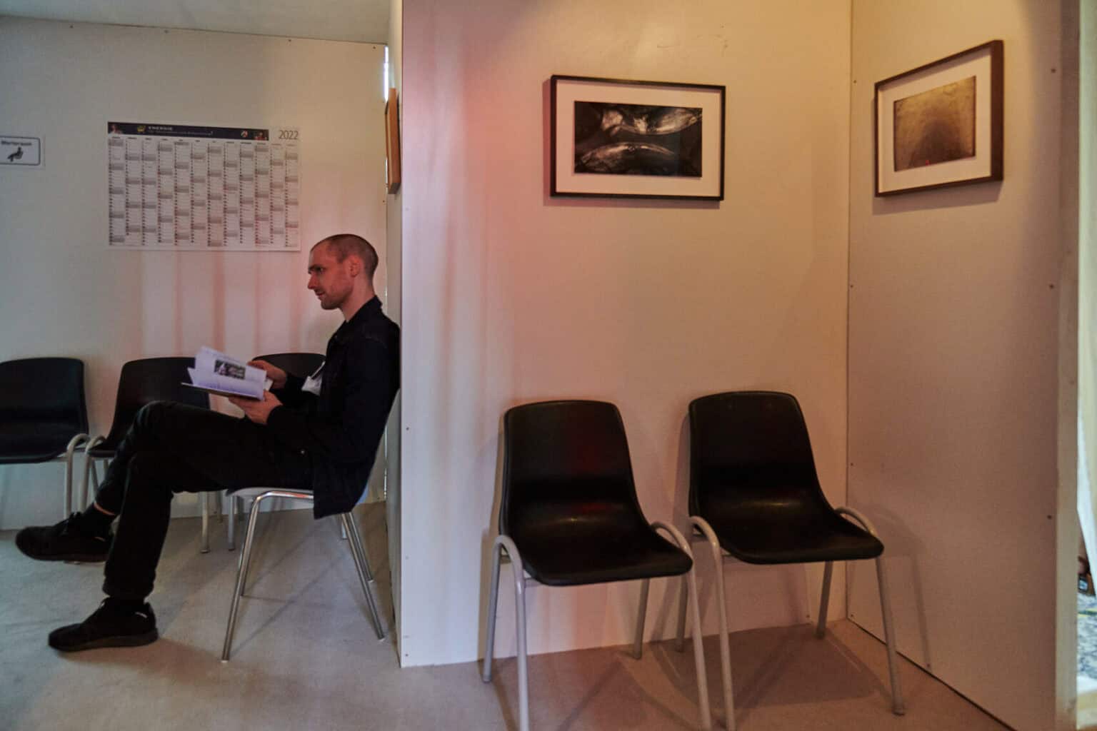 Foto: Ein Raum mit weißen Wänden und tiefer Decke. Im Raum stehen Stühle, an den Wänden hängen Kalender und gerahmt Objekte. Auf einem der Stühle sitzt ein Mensch.