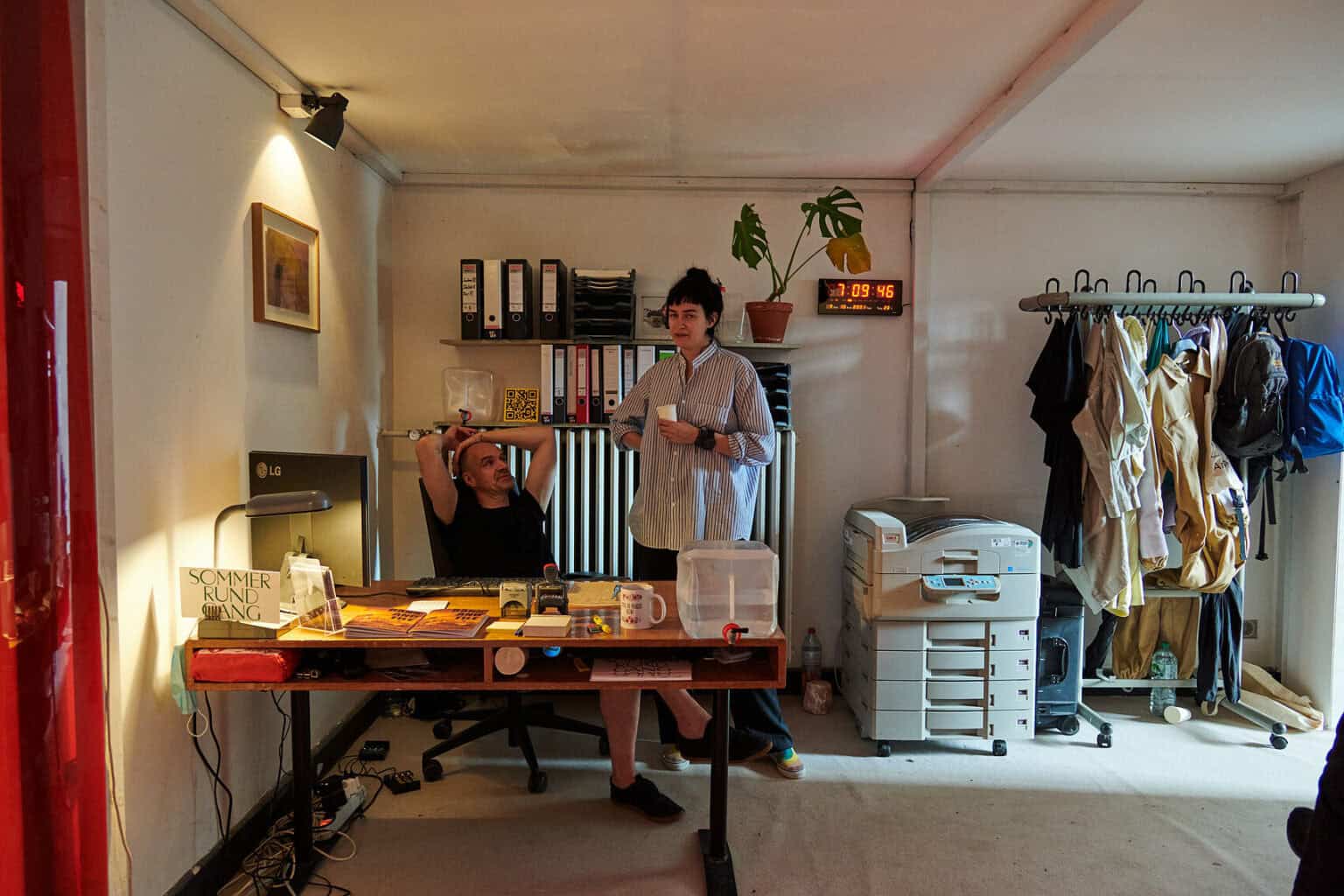 Foto: Ansicht Eines Raumes mit tiefer Decke, weißen Wänden und Büroausstattung. Darunter ein großer Drucker, Ordner, eine Tischlampe, eine Tasse, ein Bildschirm, Stempel usw. An der Wand befindet sich eine digitale Uhr und ein gerahmtes Objekt.Rechts im Bild ein Kleiderständer mit Kleidungsstücken und Taschen. Hinter dem Schreibtisch sitzen und stehen zwei Menschen.