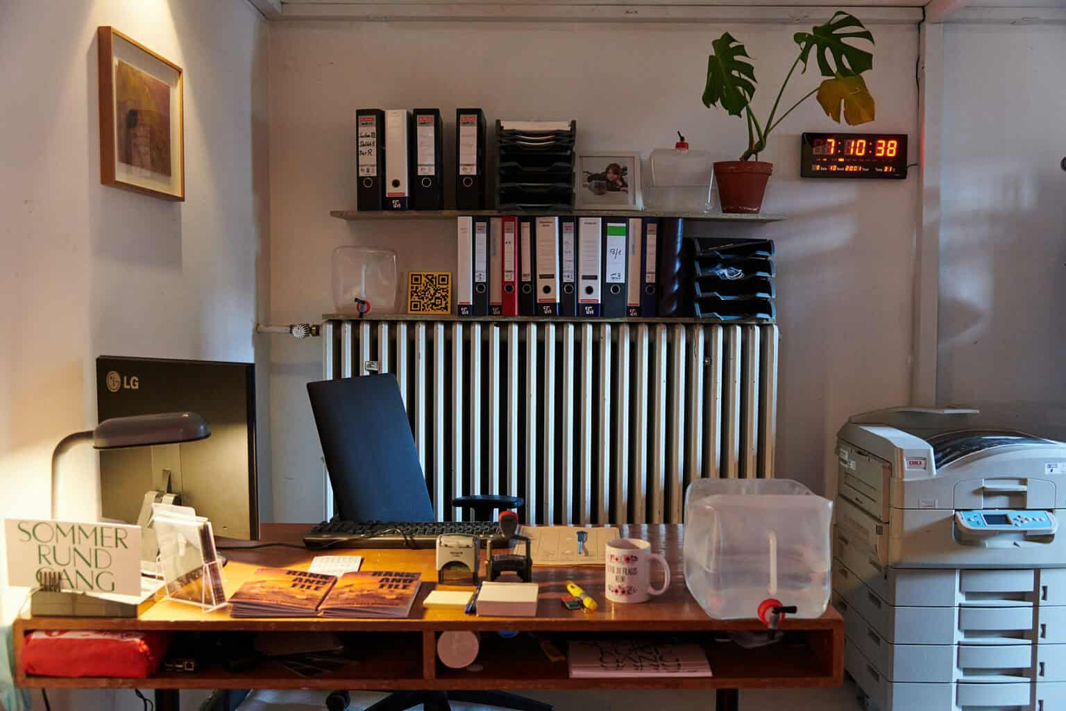 Foto: Ansicht Eines Raumes mit weißen Wänden und Büroausstattung. Darunter ein großer Drucker, Ordner, eine Tischlampe, eine Tasse, ein Bildschirm, Stempel usw. An der Wand befindet sich eine digitale Uhr und ein gerahmtes Objekt.