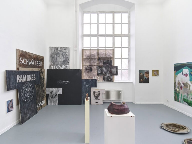 Foto: Aufnahme eines Raumes mit weißen Wänden, grauem Boden und einem großen Fenster. An den Wänden hängen und lehnen Malereien in Klein-, Mittel- und Großformaten. Auf dem Boden des Raumes und auf Sockeln befinden sich Objekte.