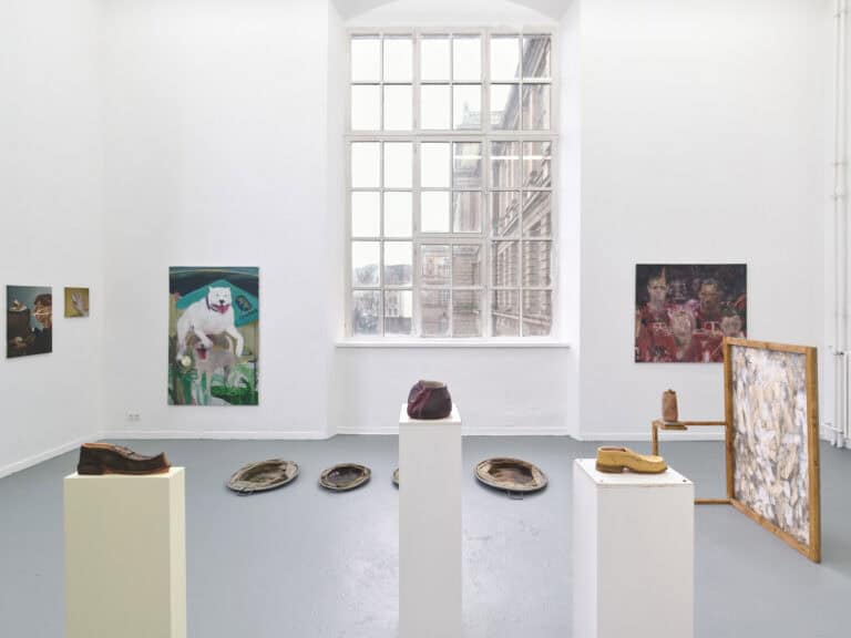 Foto: Ansicht eines Raumes mit weißen Wänden, grauem Boden und einem großen Fenster. An der Wand hängen Malereien im Klein-, Mittel- und Großformat. Auf dem Boden des Raumes und auf Sockeln befinden sich Objekte.