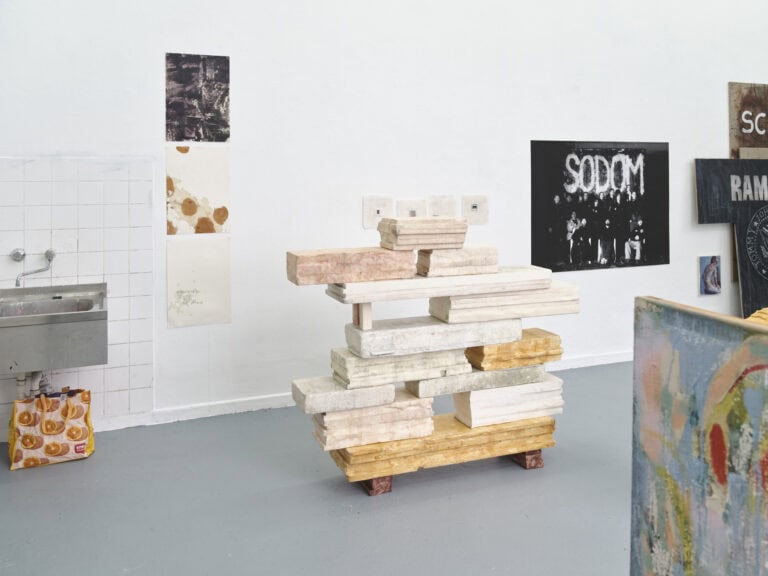 Foto: Ansicht eines Raumes mit weißen Wänden und grauem Boden. An den Wänden befinden sich flache Objekte. Auf den Boden steht eine Skulptur.