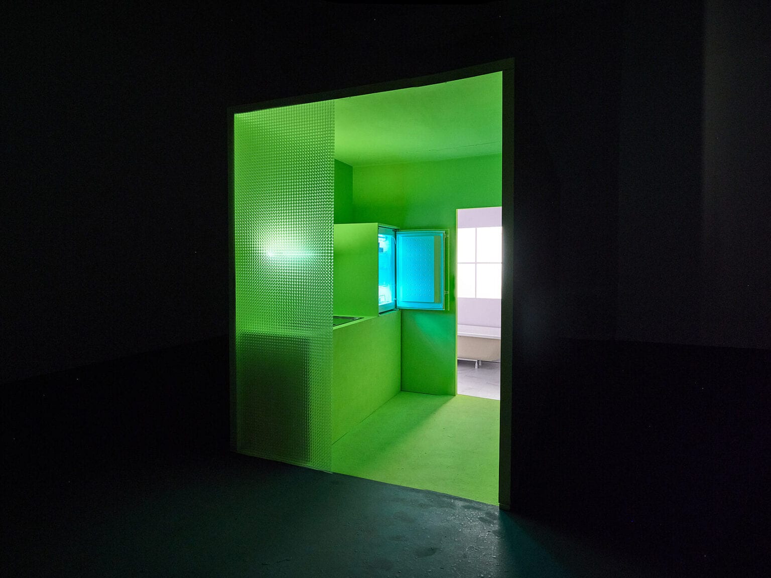 Foto: Ansicht eines dunklem Raumes mit Ausblick auf einen grün gestrichenen Raum. Innerhalb des grünen Raumes befindet sich ein grünes Objekt, aus dem ein bläuliches Licht in den Raum fällt. In einem Ausblick dahinter ist ein weißer Raum mit Fenster zu erkennen.