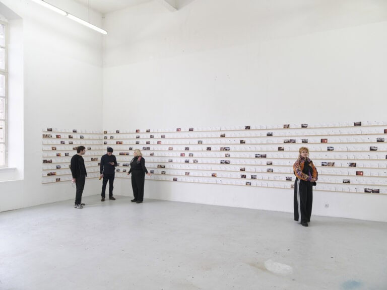 Foto: Ansicht eines Raumes mit hellgrauem Boden und einem großen Fenster. An zwei Wänden des Raumes befindet sich eine Konstruktion aus schmalen Latten an der Wand. Diese sind in mehreren Reihen in einem gleichen Abstand zueinander angebracht. Auf den Latten lehnen viele Papiere in Postkartenformat. Im Raum stehen und bewegen sich vier Menschen.