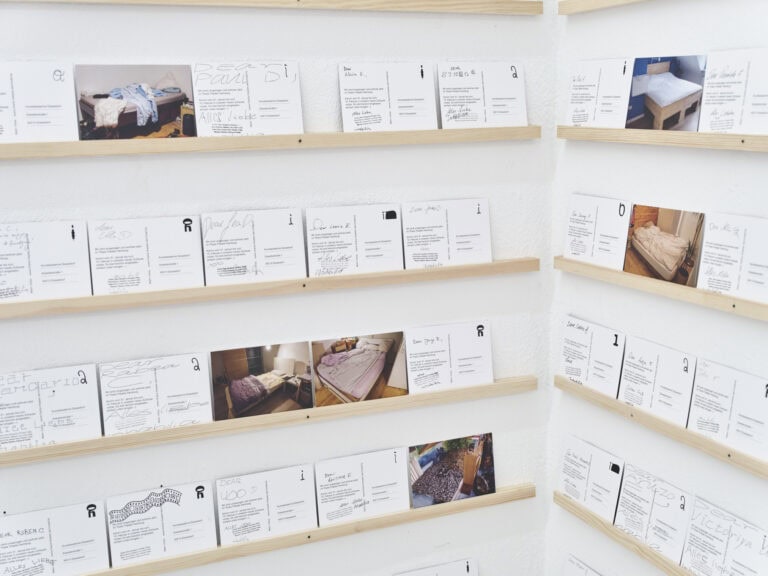 Foto: Nahaufnahme einer Raumecke mit weißen Wänden. An der Wand sind mehrere schmale Latten in gleichem Abstand zueinander angebracht, auf denen nebeneinander viele Papiere in Postkartenformat lehnen.