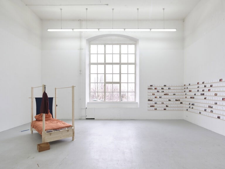 Foto: Ansicht eines Raumes mit weißen Wänden, hellgrauem Boden und einem großen Fenster. Links im Raum steht eine Skulptur. Rechts im Raum befindet sich eine Konstruktion aus schmalen Latten an der Wand. Diese sind in mehreren Reihen in einem gleichen Abstand zueinander angebracht. Auf den Latten lehnen viele Papiere in Postkartenformat.