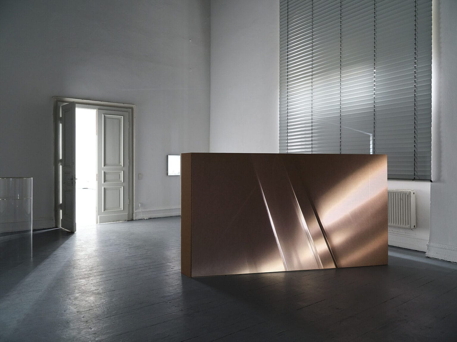 Foto: Ein abgedunkelter Raum mit weißen Wänden und grauem Boden, in den durch eine Tür Licht fällt. Die Rollläden sind heruntergelassen. Im Raum stehen und an der Wand hängen Objekte.