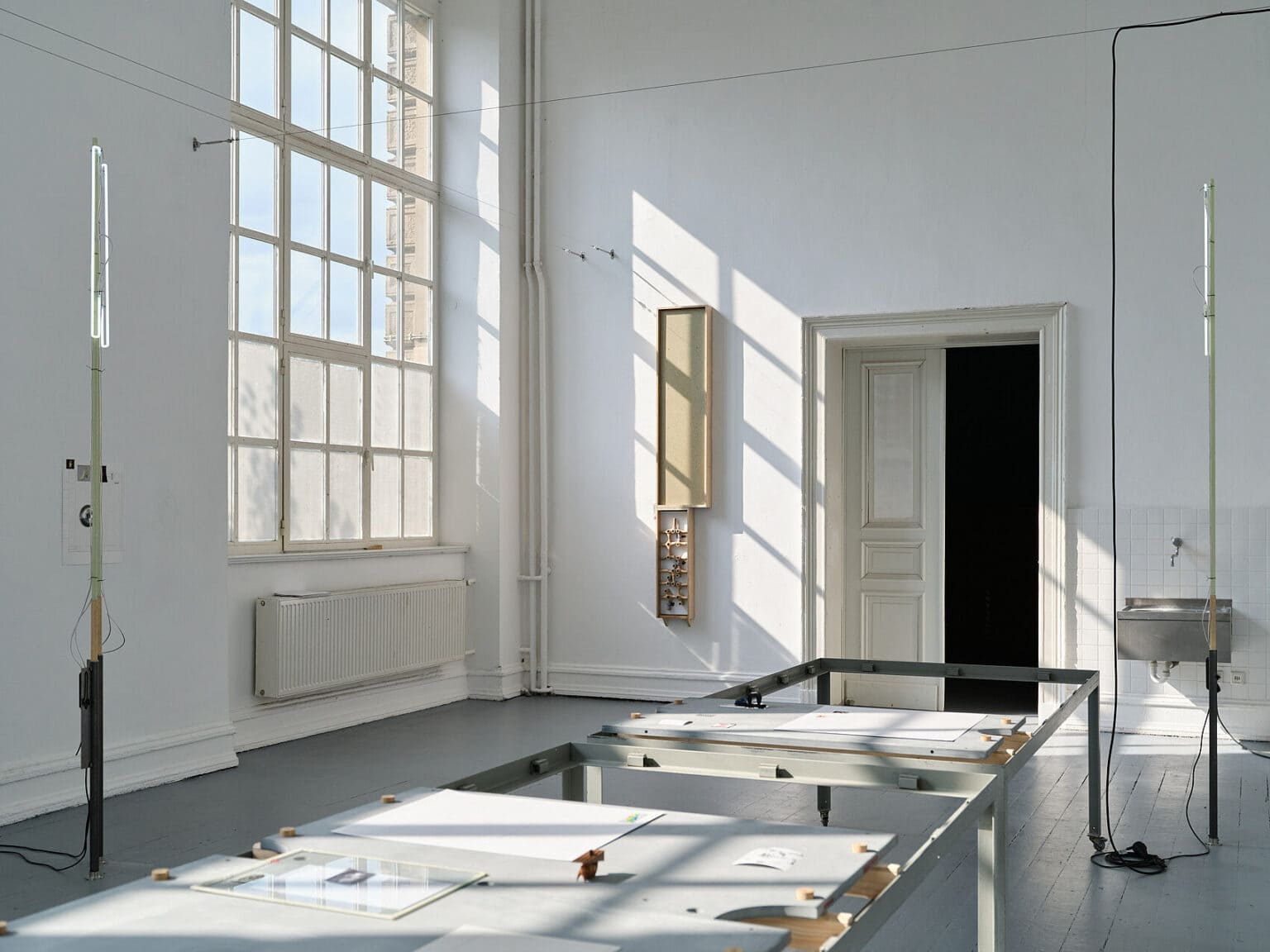 Foto: Raum mit weißen Wänden, grauem Dielenboden und einem großen Fenster, auch das Licht in den Raum Fällt. Im Raum und an den Wänden befinden sich viele Objekte und in der oberen Hälfte des Raumes sind Drahtseile gespannt.