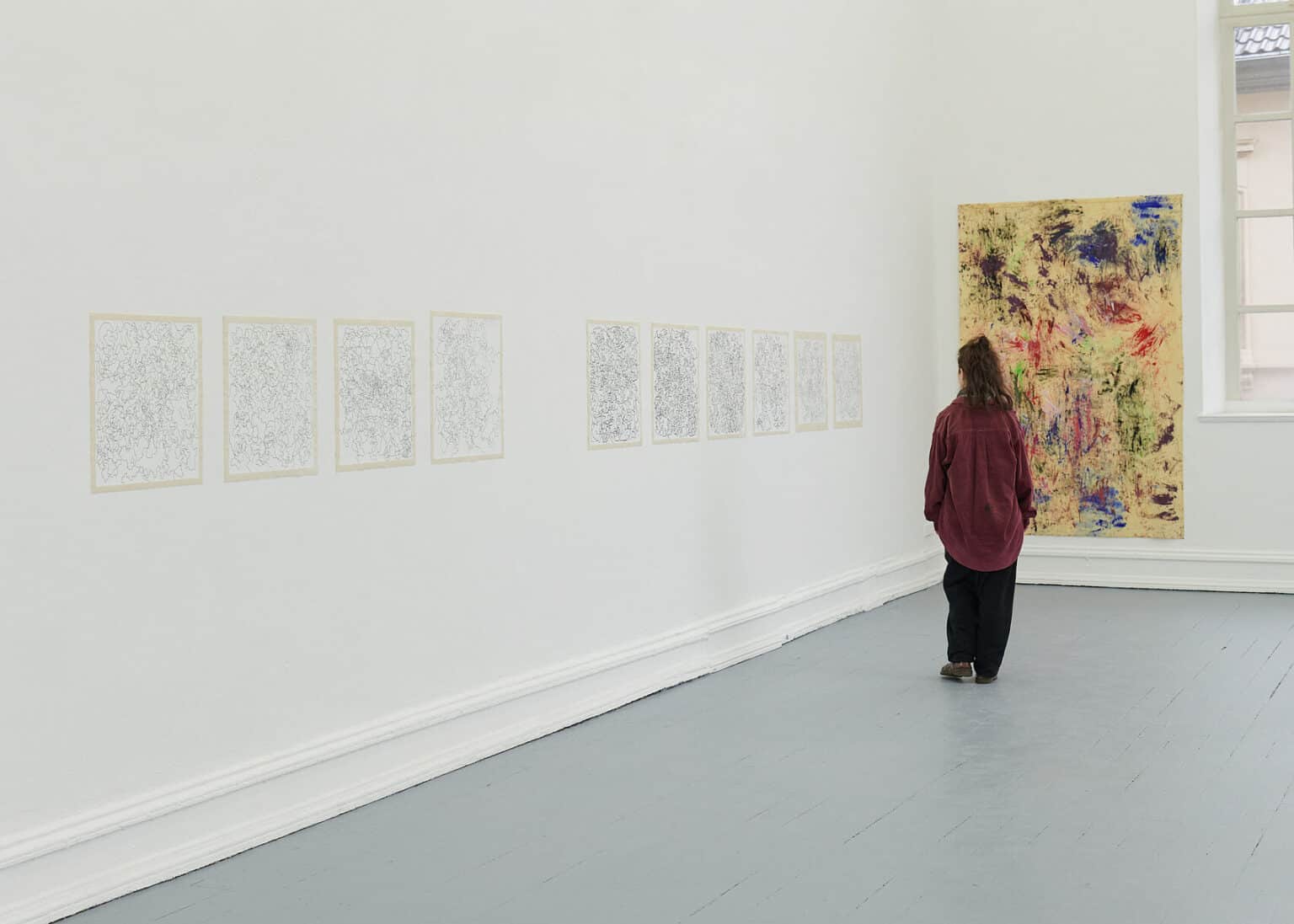 Foto: Ansicht eines Raumes mit weißen Wänden, breiten Fußleisten, grauem Dielenboden und einem Fenster. An einer der Wände hängen, in einer Linie und mit Tape fixierte, zehn Papierarbeiten. An einer der Wände hängt eine großformatige Leinwand. Durch den Raum bewegt sich eine Person.