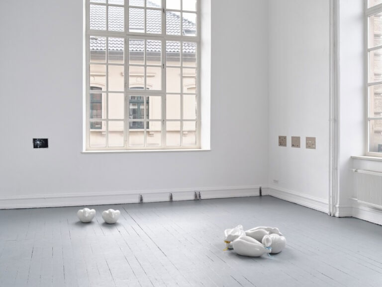 Foto: Ansicht eines Raumes mit weißen Wänden, grauem Boden und zwei Fenstern. An den Wänden befinden sich kleinformatige Objekte. Am Boden des Raumes und an der Wand lehnen weitere Objekte.