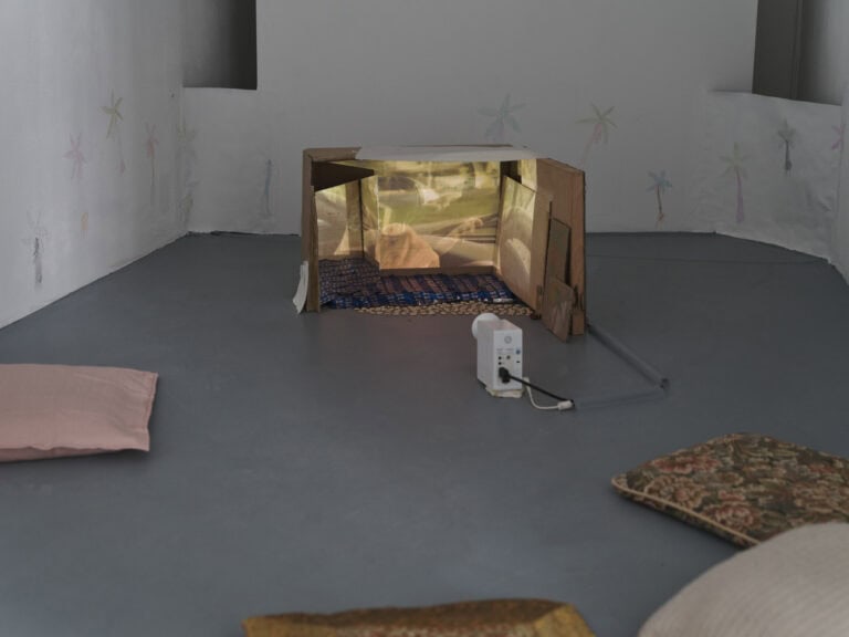 Foto: Nahaufnahme eines Objektes im Raum. Die Wände sind weiß und der Boden grau, Kissen liegen auf dem Boden. Tiefer im Raum steht ein Objekt auf dem Boden, dass von einem kleinen Projektor angeleuchtet wird. Die Wände sind mit blassen Zeichnungen versehen.