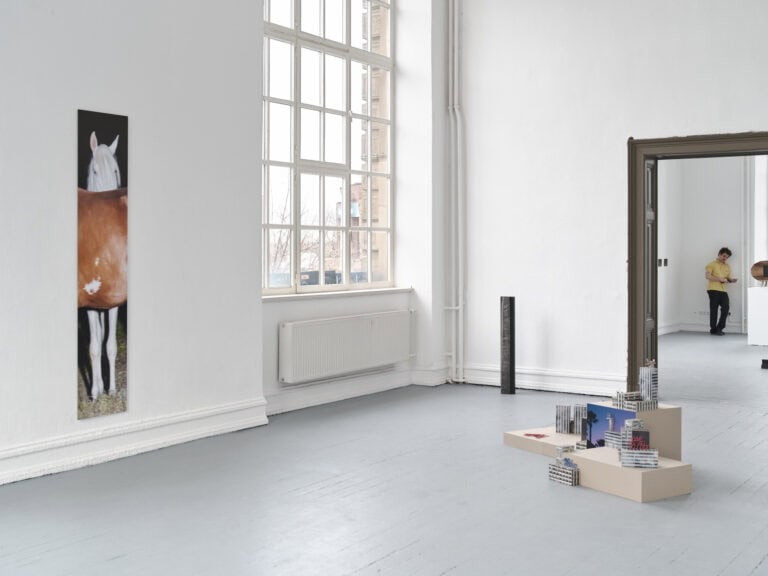 Foto: Ansicht eines Raumes mit weißen Wänden, grauem Boden und einem großen Fenster. An der Wand befinden sich ein schmales Objekt. Im Raum verteilt stehen Objekte direkt auf dem Boden oder auf Sockeln. Im Hintergrund steht ein Mensch, der sich an die Wand lehnt.