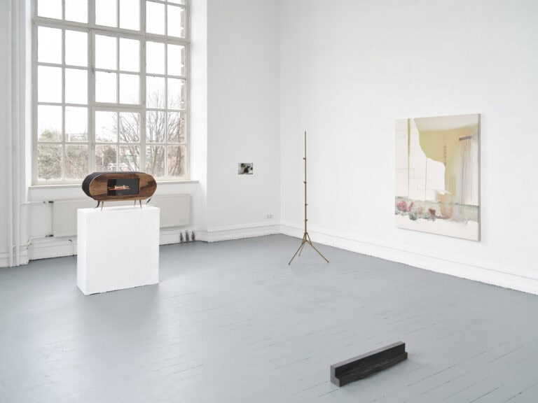 Foto: Ansicht eines Raumes mit weißen Wänden, grauem Boden und einem großen Fenster. An der Wand befinden sich Objekte in Klein- und Großformate. Im Raum befinden sich auf dem Boden stehen, an der Wand lehnend und auf einem Sockel installiert weitere Objekte.