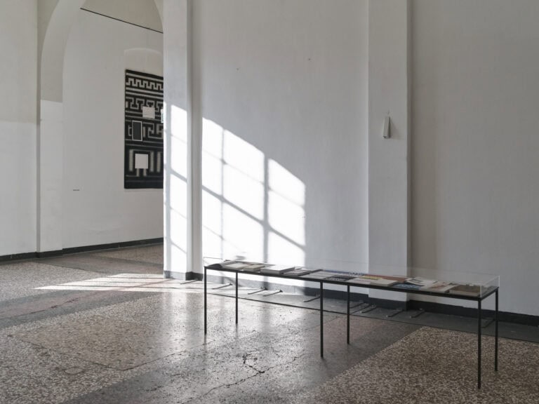 Foto: Ansicht eines Raumes mit weißen Wänden und mehrfarbigen Steinboden. Im Vordergrund eine Vitrine gefüllt mit Büchern und Flyer. Der Schatten eines Bogenfenster fällt in den Raum. Im Hintergrund ein flaches, gemustertes Panel an der Wand.