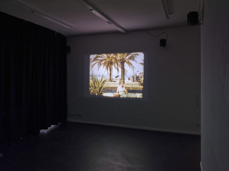 Foto: Ansicht eines abgedunkelter Raumes mit einem dunklen Vorhang. Auf die Wand wird eine Projektion geworfen. Oben an der Wand befinden sich Lautsprecher.