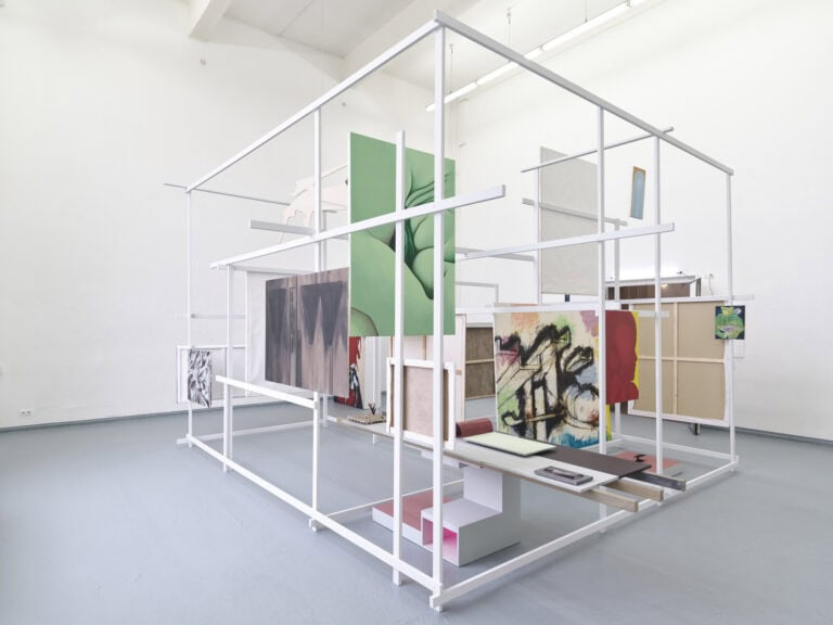 Foto: Ansicht eines Raumes mit weißen Wänden und grauem Boden. Im Raum befindet sich eine verschachtelte rechteckige Konstruktion aus Holzlatten. An die Holzlatten sind Leinwände und Platten angebracht. Auf den Platten liegen Objekte.