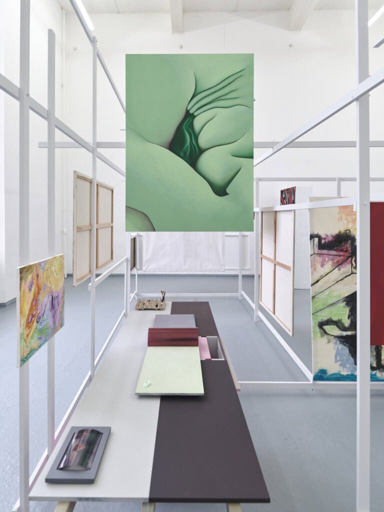 Foto: Ansicht eines Raumes mit weißen Wänden und grauem Boden. Im Raum befindet sich eine verschachtelte Konstruktion aus Holzlatten. An die Holzlatten sind Leinwände und Platten angebracht. Auf den farbigen Platten liegen Objekte.