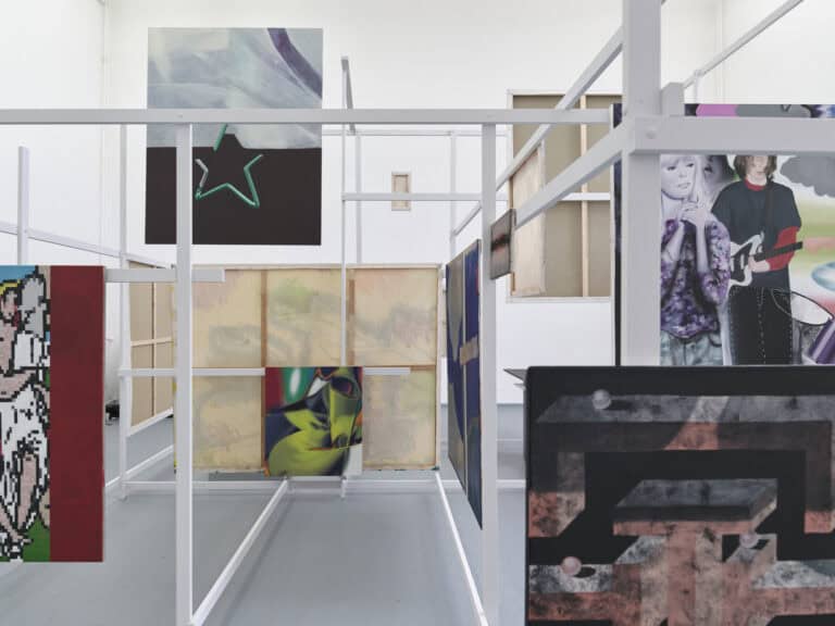 Foto: Ansicht eines Raumes mit weißen Wänden und grauem Boden. Im Raum befindet sich eine verschachtelte Konstruktion aus Holzlatten. An die Holzlatten sind Leinwände angebracht.