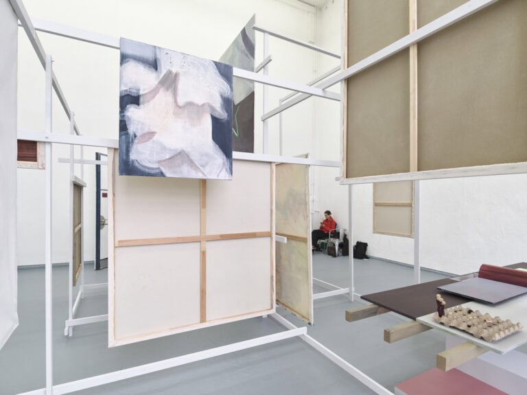 Foto: Ansicht eines Raumes mit weißen Wänden und grauem Boden. Im Raum befindet sich eine verschachtelte Konstruktion aus Holzlatten. An Die Holzlatten sind Leinwände und Platten angebracht. Auf den Platten liegen Objekte. Im Hintergrund sitzt ein Mensch auf einem Stuhl.