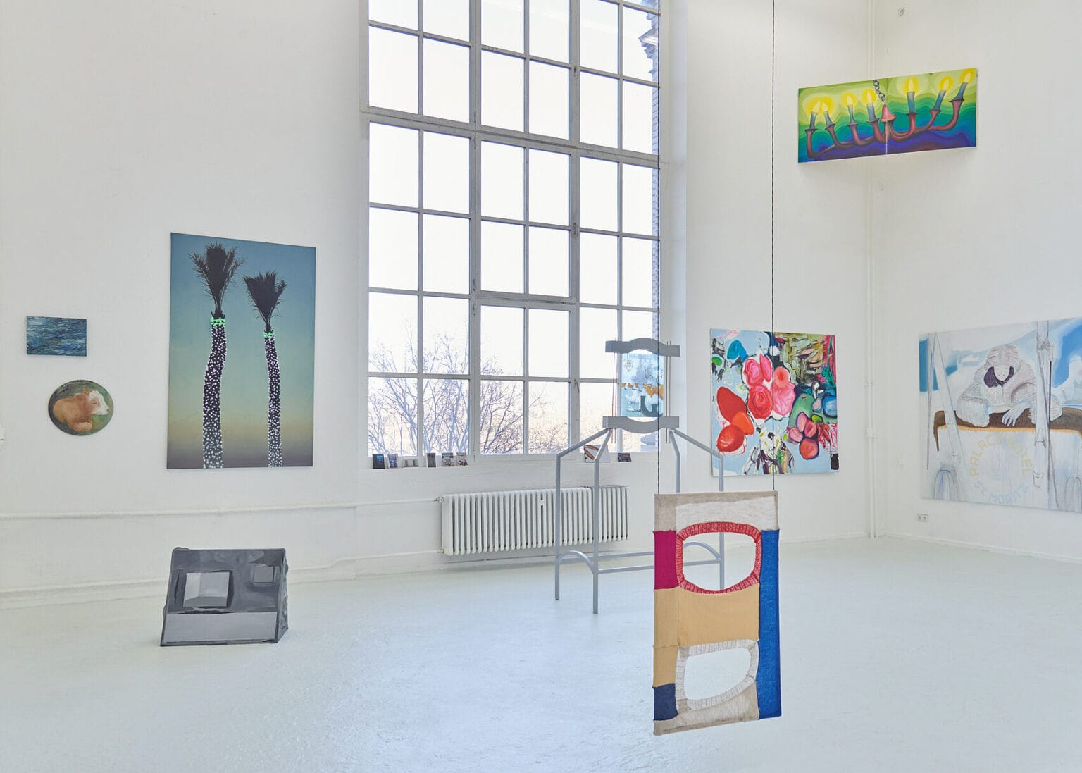 Foto: Ansicht eines Raumes mit weißen Wänden, gräulichem Boden und einem großen Bogenfenster. An den Wänden hängen, auf unterschiedlichen Höhen, zum teil auch übereinander und quer in den Ecken des Raumes, Malereien im Klein- und Großformat. Im Raum befinden sich Objekte. Manche von ihnen stehen auf dem Boden, andere hängen von der Decke.