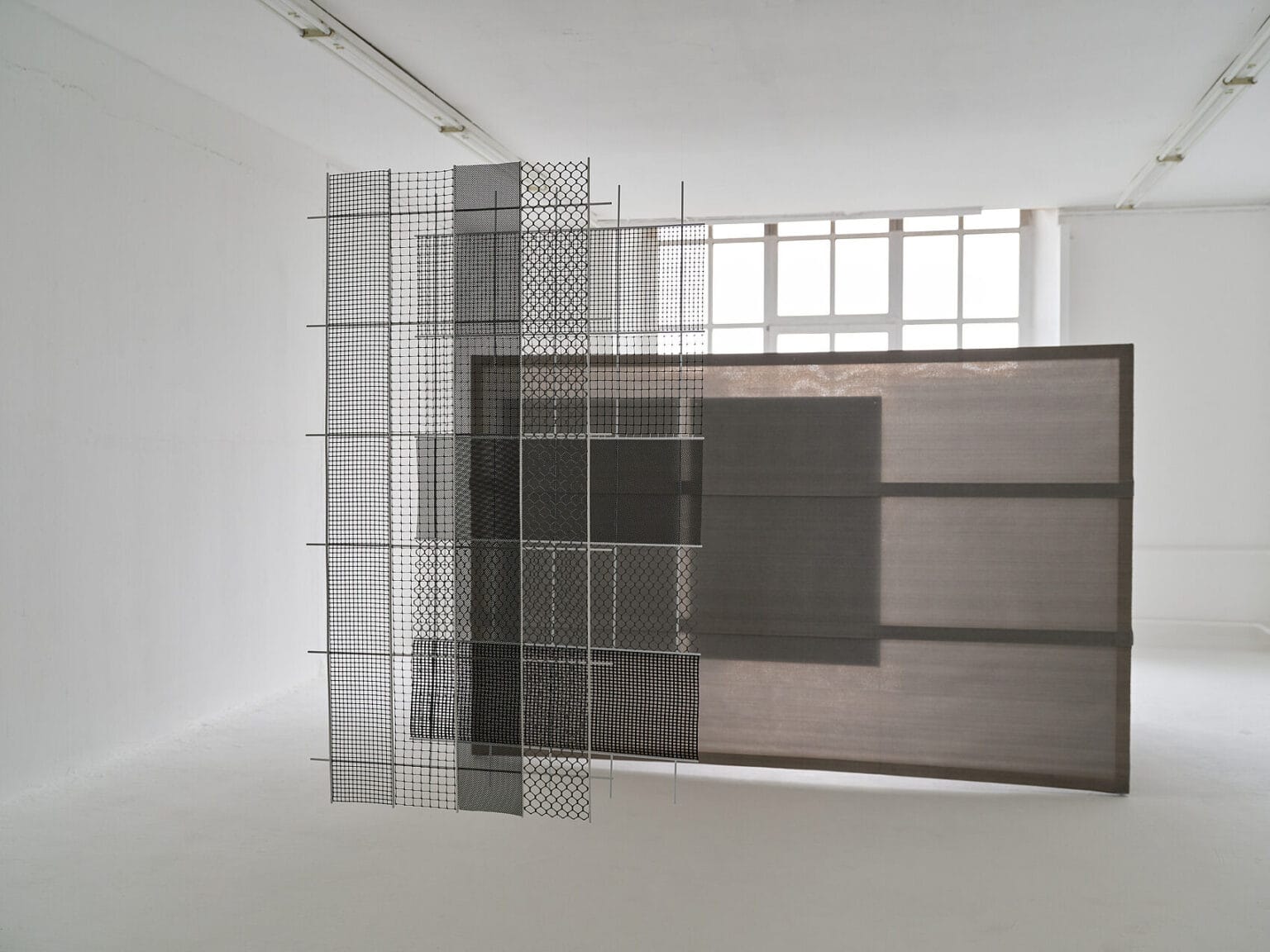 Foto: Ansicht eines Raumes mit weißen Wänden, weißem Boden und einem Fenster. Mittig im Raum befinden sich mehrere, zum teil transparente und gitterartige Objekte, die hintereinander stehen.