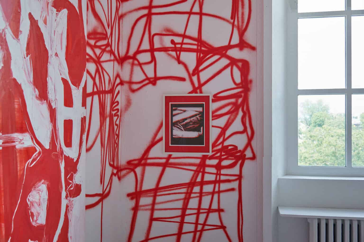 Foto: Ein Raum mit weißen Wänden und grauem Boden und Fenster. Die Wände sind mit einem Muster aus roten Linien besprüht. Auf einer dieser Wände hängen Arbeiten.