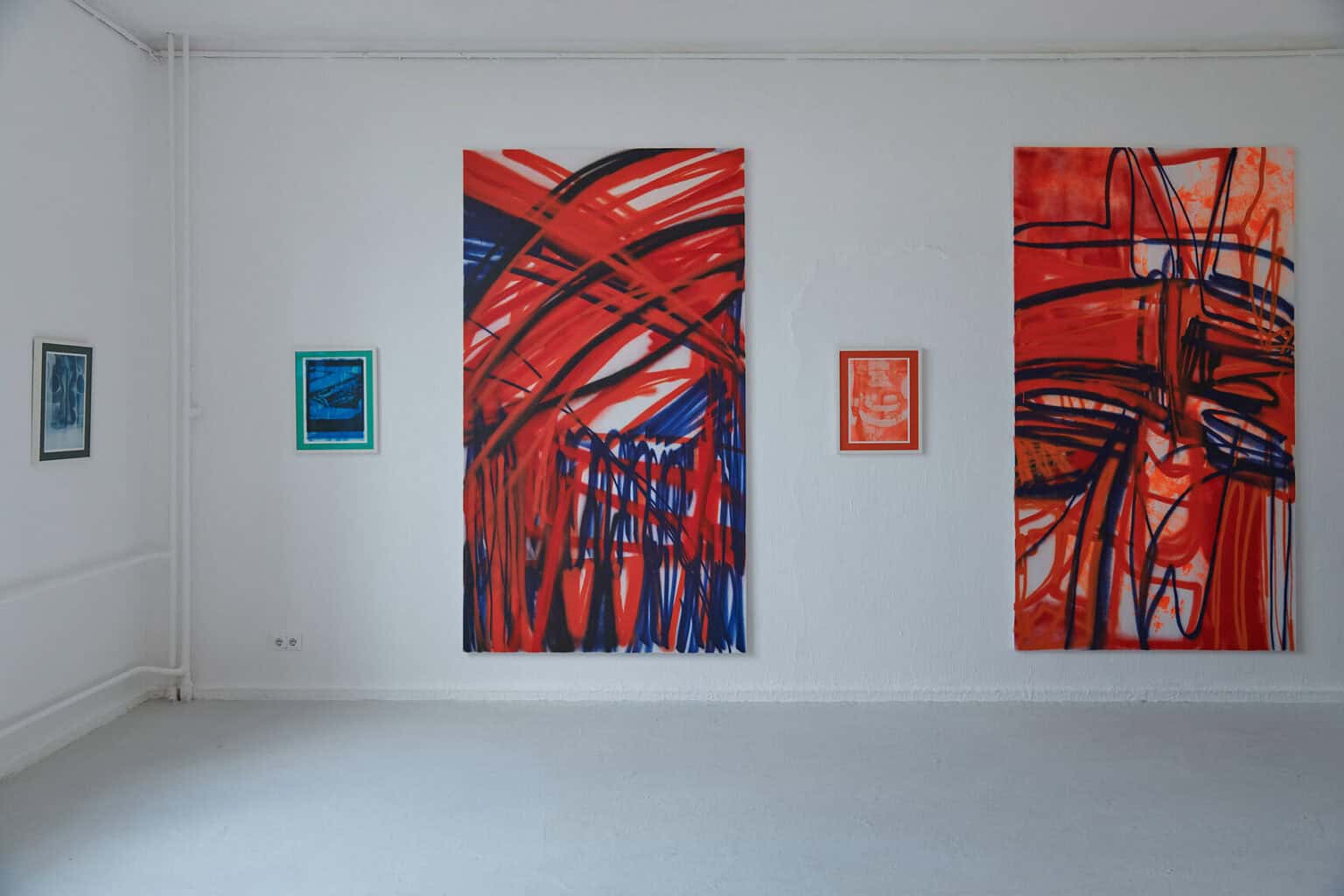 Foto: Ein Raum mit weißen Wänden und grauem Boden. An den Wänden hängen, abwechselnd, klein- und großformatige Arbeiten an der Wand.