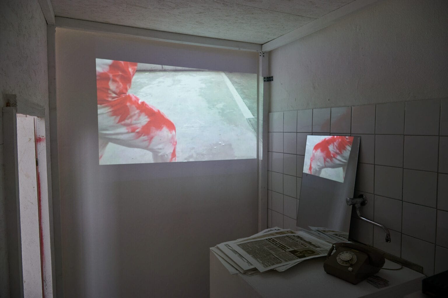 Foto: Ein kleiner, halbdunkler Raum mit weißen Wänden und Fliesen. Im Raum befinden sich eine große und eine kleine Projektion, die ein ähnliches Motiv zeigen. Vor einer der Projektionen liegt ein Zettelhaufen, daneben steht ein Telefon.