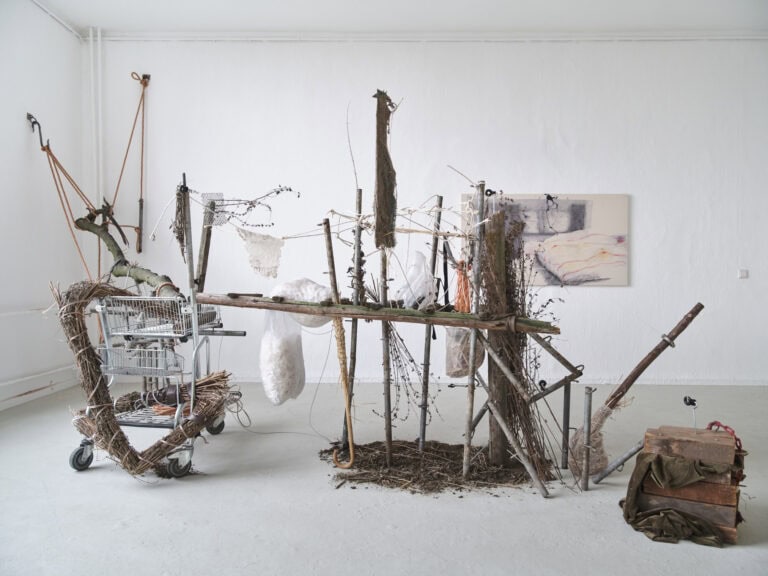 Foto: Ansicht eines Raumes mit weißen Wänden und grauem Boden. Im Raum befindet sich ein Objekt aus vielen unterschiedlichen Materialien, die zum Teil auf dem Boden stehen und liegen, ineinander gesteckt und gebunden sind und von der Wand hängen. An der Wand im Hintergrund befindet sich eine Malerei.