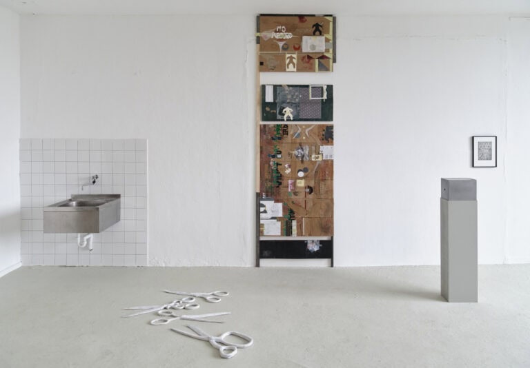 Foto: Ansicht eines Raumes mit weißen Wänden und grauem Boden. Links im Bild ein von Kacheln umrahmtes Waschbecken. An den Wänden befinden sich zwei flache Objekte. Auf dem Boden liegen und stehen Skulpturen.