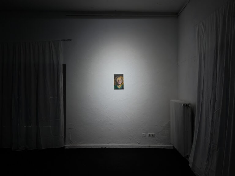 Foto: Ansicht eines abgedunkelter Raumes. Vor der weißen Wand hängen links und rechts im Bild zwei weiße Textilien, die Falten werfen. An der Wand hängt eine kleinformatige Malerei, die von einem Lichtkegel angestrahlt wird.