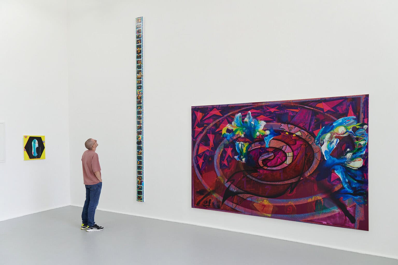 Foto: Ein Raum mit weißen Wänden und grauem Boden. An der Wand befinden sich Objekte aus unterschiedlichen Materialien und in unterschiedlichen Formaten. Eines von ihnen ist sehr schmal und hoch. Ein Mensch steht vor diesem Gegenstand und blickt hoch.