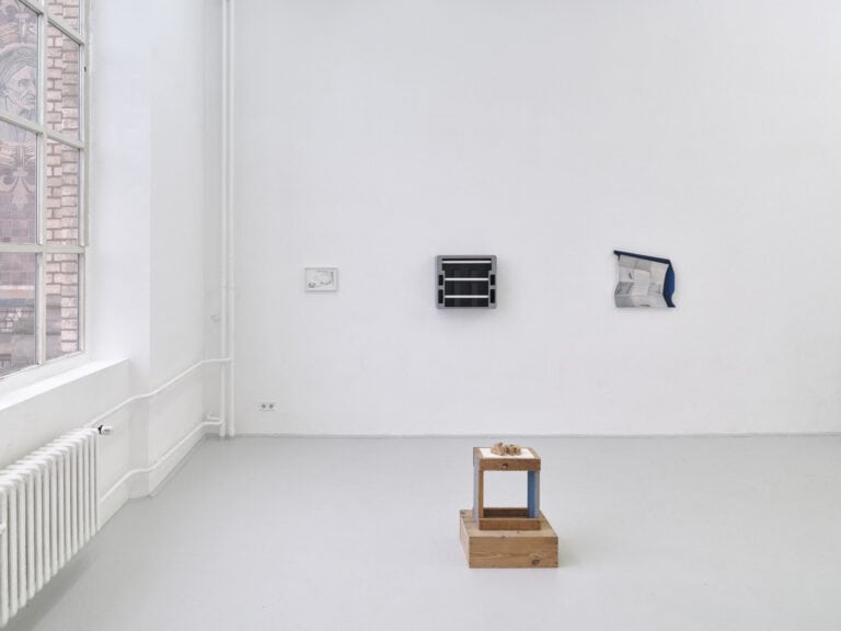 Foto: Ansicht eines Raumes mit weißen Wänden, grauem Boden und großem Fenster. An den Wänden befinden sich Objekte im Klein- und Mittelformat. Im Raum befinden sich weitere Objekte, die zum Teil auf Sockeln stehen.